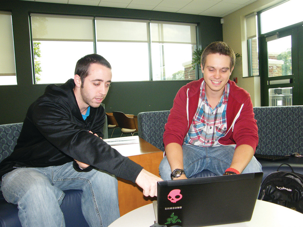 IN THE GAME: Corey King, 22, at left, and John Groh, 20, work on their game, “Parkourasaur,” in the student lounge of New England Institute of Technology, where they are students. The pair, who met at the school in 2010, hope to raise $7,000 to fund the game and release it later this year.